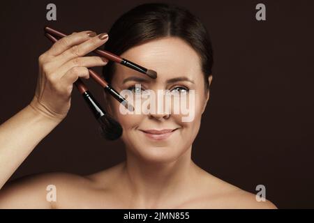 Donna di mezza età con spazzole per il trucco su sfondo marrone Foto Stock