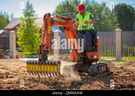 Un uomo su un miniescavatore livella un pezzo di terra, allenta il terreno Foto Stock