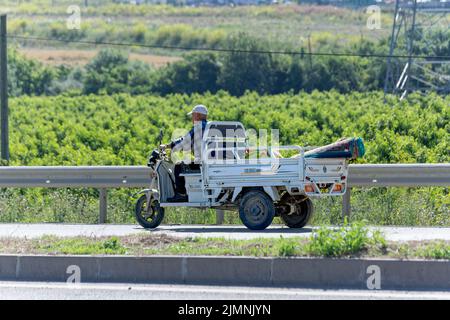 Il vecchio uomo trasporta il tappeto con il pick-up elettrico a tre ruote sull'autostrada. Foto Stock