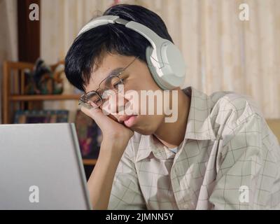 Annoiato studente addormentato indossando cuffie stanco di imparare a fare i compiti seduti mezzo sonno allo schermo del laptop nella sua stanza. Ragazzo adolescente triste che studia p Foto Stock