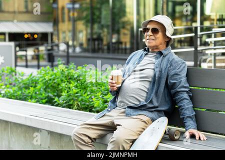 Uomo anziano con un longboard seduto sulla panchina e bere caffè in un parco cittadino Foto Stock