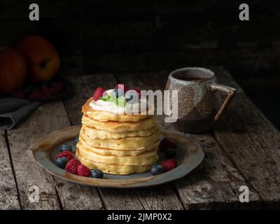 Pancake con crema alla vaniglia, mirtilli e lamponi. Moody scuro vecchio sfondo rustico in legno. Foto Stock