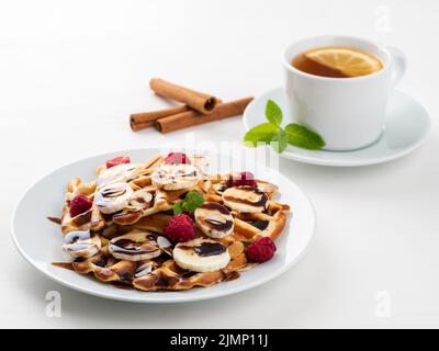 Waffle belgi con lamponi, banana, sciroppo al cioccolato. Colazione con tè su sfondo bianco, vista laterale Foto Stock