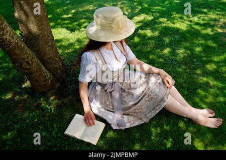 Una donna in un bel vestito tradizionale bavarese dirndl (o Tracht) leggendo un libro sul prato verde in una giornata estiva soleggiata (Monaco, Baviera, tedesco Foto Stock