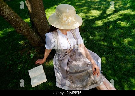 Una donna in un bel vestito tradizionale bavarese dirndl (o Tracht) leggendo un libro sul prato verde in una giornata estiva soleggiata (Monaco, Baviera, tedesco Foto Stock