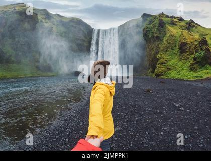 Donna asiatica in giacca gialla che tiene le mani con la coppia di fronte alla cascata di Skógafoss che scorre sulla scogliera in estate nel sud dell'Islanda Foto Stock