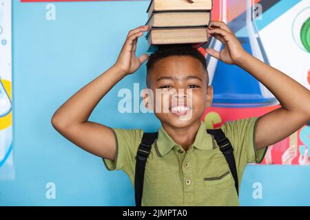 Ritratto di sorridente scolaro afroamericano elementare che accatastano i libri sulla testa contro il muro Foto Stock