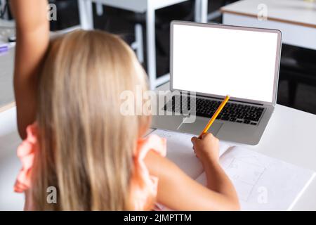 Vista posteriore di una studentessa elementare caucasica che utilizza un computer portatile mentre si siede con la mano sollevata in classe Foto Stock