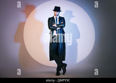 Ritratto a tutta lunghezza di un uomo bello e sicuro in cappotto nero e cappello con braccia incrociate in posa in studio sotto i riflettori. Foto Stock