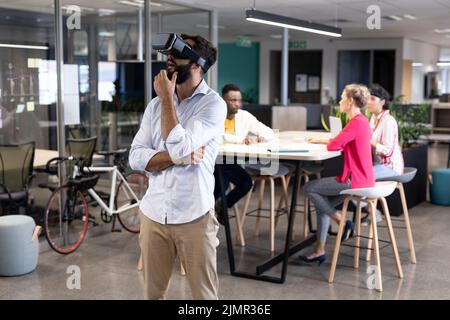 Un uomo d'affari caucasico attento che indossa un simulatore di realtà virtuale contro colleghi biraciali Foto Stock