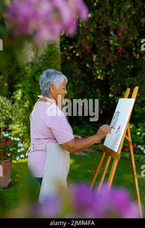 Vista laterale di sorridente donna anziana biraciale con pittura corta dei capelli su tela contro le piante nel cortile Foto Stock