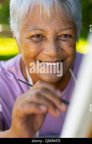 Primo piano ritratto di sorridente biraciale Senior Woman pittura con pennello su tela in cantiere Foto Stock