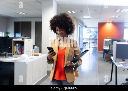 Sorridente donna d'affari ispanica che cammina mentre si utilizza lo smartphone sul luogo di lavoro moderno Foto Stock