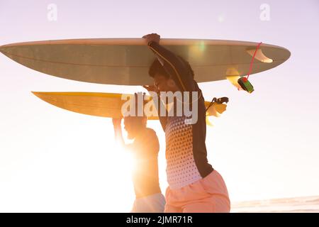 Coppia afroamericana che porta tavole da surf sulla spiaggia contro il cielo limpido durante il tramonto Foto Stock