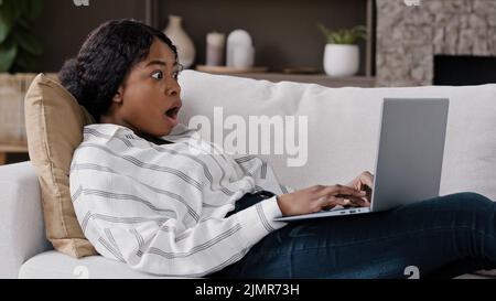 Ragazza africana freelancer donna biraciale americana sdraiata sul divano a casa guardando il laptop lettura inaspettata cattive notizie gossip ricezione notifica Foto Stock