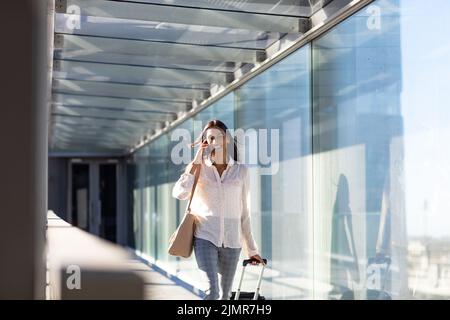 Sorridente giovane donna d'affari biraciale che parla al telefono camminando con i bagagli nel corridoio dell'aeroporto Foto Stock