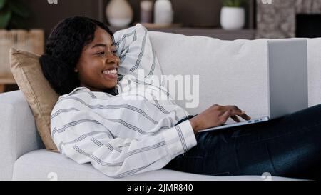 Sorridente donna africana studente ragazza freelance utente sdraiato sul divano utilizzando il portatile digitando messaggio online chat con gli amici felice chattare sul computer Foto Stock