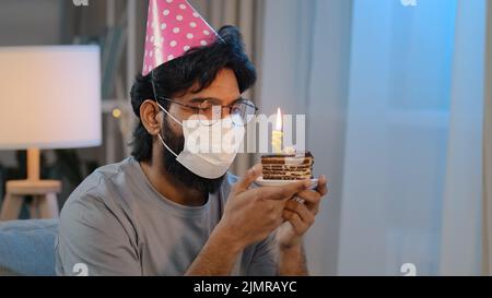 Ritratto arabo malato malato indiano bearded triste uomo in maschera medica seduta da solo a casa indossa festive compleanno rosa cappello e bicchieri tiene cupcake Foto Stock