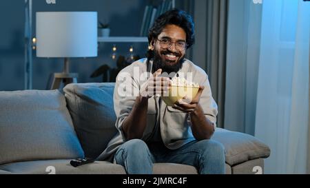 Uomo bearded in occhiali indiano maschio bachelor dopo il lavoro si siede sul divano nel soggiorno scuro accende la TV con telecomando che sceglie il canale di guardare Foto Stock
