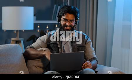 L'uomo bearded divertente indiano arabo indossa cuffie senza fili che hanno divertimento ascoltare musica con il laptop a casa seduta divano sera tardi Foto Stock