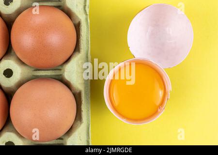 Confezione di uova marroni con un uovo aperto rotto con tuorlo crudo e guscio su sfondo giallo Foto Stock