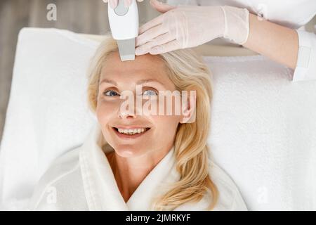 Estetician fare la pulizia ultrasonica del viso per la donna abbastanza felice di mezza età, primo piano Foto Stock