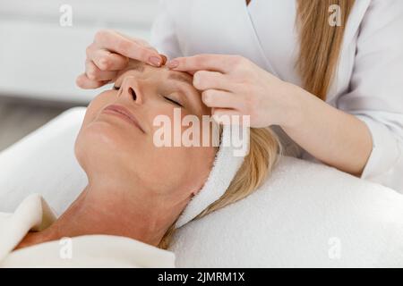 La cosmetologa femminile sta facendo il massaggio facciale per la donna matura bionda bella in armadietto di bellezza della luce Foto Stock