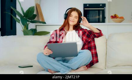 La giovane ragazza spensierata con il laptop in posa di lotus ascolta la musica preferita ad alta voce ama la traccia audio teenage redhead donna caucasica indossa le cuffie Foto Stock