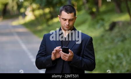 Uomo caucasico adulto serio stand all'aperto in parco indossa abito nero formale tenere telefono cellulare scorrimento pagine di scorrimento sullo schermo dello smartphone Foto Stock