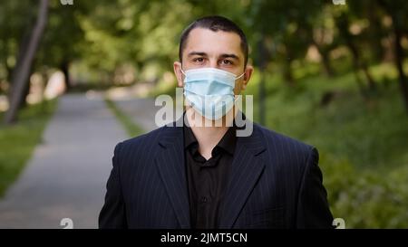 Uomo d'affari caucasico stanco toglie maschera medica respiratore protettivo uomo esausto si leva in piedi nel parco della città sente la libertà fa respiro profondo Foto Stock