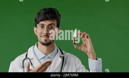 Arabo giovane maschio ispanico medico indiano in cappotto bianco isolato in studio verde raccomandare a fotocamera spray nasale medicina rimedio liquido per il naso Foto Stock