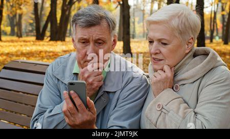 Sconvolto anziano coppia sposata sedersi in panchina in autunno parco leggere cattive notizie sullo smartphone preoccupato persone anziane guardare schermo telefono triste famiglia sensazione scioccante Foto Stock
