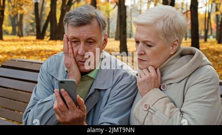 Sconvolto anziano sposato coppia seduta su panchina in autunno parco leggere cattive notizie su smartphone preoccupato persone anziane guardare schermo telefono triste famiglia sensazione Foto Stock