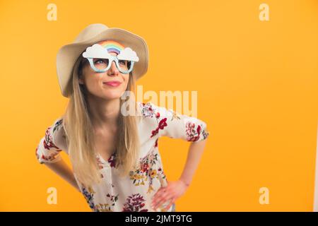 Giovane donna bianca bionda in estate cappello e occhiali a forma di arcobaleno in piedi con le mani sui fianchi che si appoggiano vicino alla fotocamera guardando a distanza. Foto studio. Foto di alta qualità Foto Stock