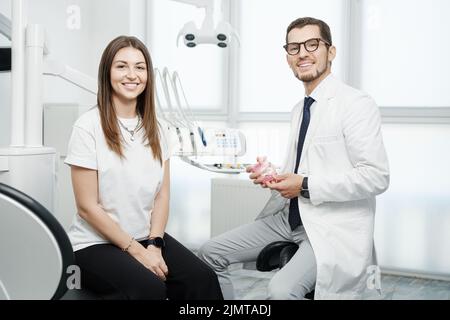 Buon dentista maschile seduto vicino al riunito con paziente caucasico femmina con un sorriso guardando la macchina fotografica Foto Stock