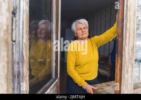 Vecchia donna caucasica in pensione grigio-capelli fissando fuori la porta aperta del balcone in una giornata di inverno. Foto di alta qualità Foto Stock