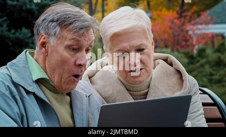 Scioccato sconvolto anziano sposato coppia partner guardando il notebook schermo sedersi in autunno parco deluso vecchia donna leggere cattive notizie preoccupata famiglia matura Foto Stock