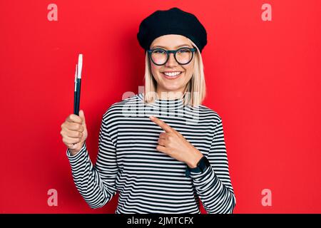 Bella donna bionda che indossa il look francese con beretto che tiene pennelli pittore sorridente puntamento felice con mano e dito Foto Stock