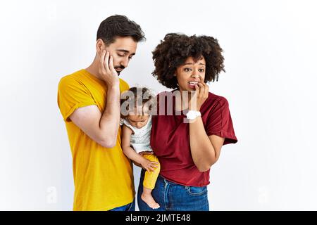 Giovane famiglia interrazziale di madre nera e padre ispanico con figlia in cerca di stress e nervoso con le mani sulla bocca morsi unghie. Ansia prob Foto Stock