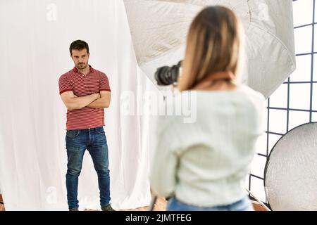 Donna fotografo che parla immagini di uomo in posa come modello presso lo studio di fotografia scettico e nervoso, disapprovando l'espressione sul viso con braccio incrociato Foto Stock