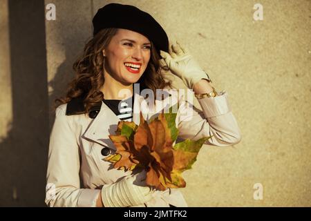 Ciao settembre. Giovane donna sorridente in trench beige con foglie di colore giallo autunno fuori sulla strada della città in autunno. Foto Stock