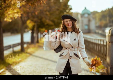 Ciao novembre. Donna alla moda sorridente in trench beige con borse shopping e foglie di giallo autunno fuori sulla strada della città in autunno. Foto Stock