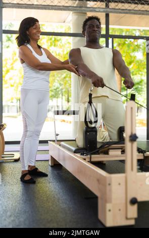 Istruttore ispanico focalizzato che aiuta l'uomo afroamericano praticando pilates. Pullman individuale Foto Stock
