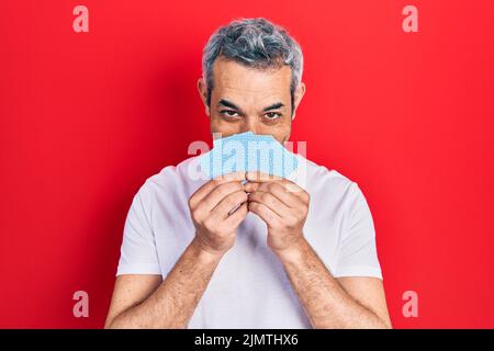 Bell'uomo di mezza età con i capelli grigi coprendo il viso con le carte sorridenti con un sorriso felice e fresco sul viso. Mostrando i denti. Foto Stock