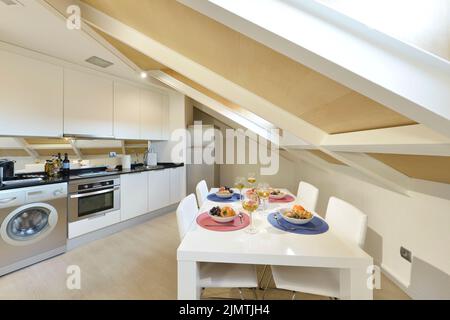 Sala da pranzo con tavolo in legno bianco con bicchieri di vino bianco e un sacco di frutta assortita, sedie rivestite in pelle bianca accanto alla cucina open space Foto Stock