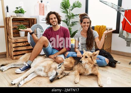 Giovane coppia ispanica che fa la lavanderia con cani sorridenti allegri che presentano e puntano con la palma di mano guardando la macchina fotografica. Foto Stock