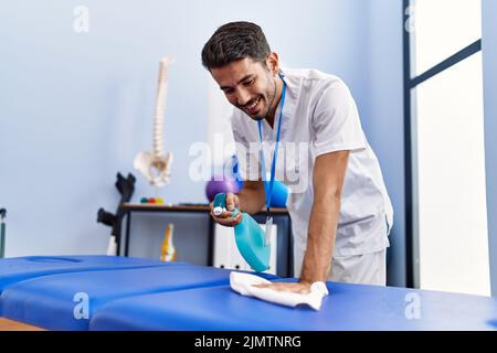 Giovane uomo ispanico che indossa fisioterapista uniforme disinfezione tavolo da massaggio alla clinica di riabilitazione Foto Stock