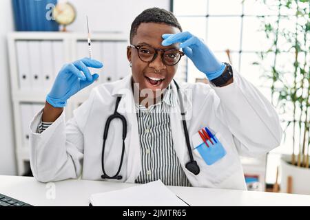 Giovane medico africano che tiene la siringa all'ospedale molto felice e sorridente guardando lontano con mano sopra la testa. Concetto di ricerca. Foto Stock