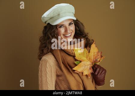 Salve settembre. Ritratto di donna sorridente di mezza età in sciarpa con guanti in pelle e foglie di acero giallo autunno isolato su backgroun bronzo Foto Stock