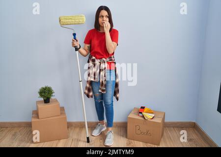 Giovane ispanica donna pittura pareti casa con rullo di vernice cercando stressato e nervoso con le mani sulla bocca morsi unghie. Problema di ansia. Foto Stock